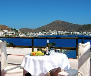 Una mesa con frutero, botella y vaso, con vistas a un pueblo costero con barcos y una montaña al fondo.