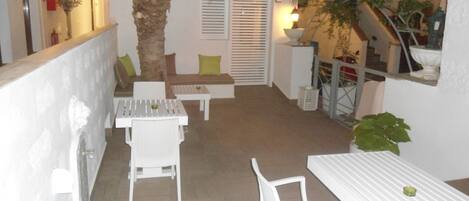 Un patio con mobiliario blanco, que incluye una mesa y sillas, un rincón de estar pequeño con cojines y una palmera.