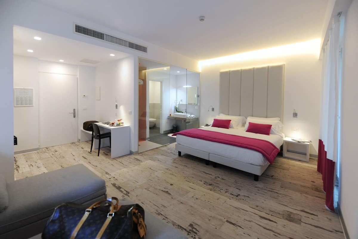 Een moderne hotelkamer met een bed, bureau, stoel, een kleine badkamer en een houten vloer.