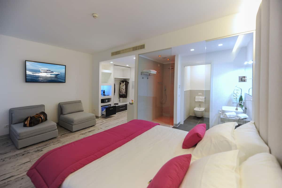 Een moderne hotelkamer met een groot bed, een zithoek met een bank en fauteuil, een inloopkast en een badkamer met glazen douchecabine.