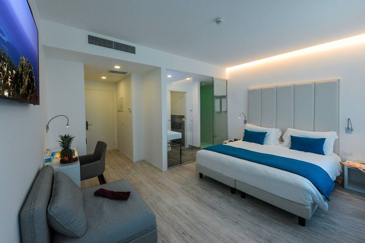 Een moderne hotelkamer met een groot bed, een sofa, een kleine tafel met een ananas en fruit, en een badkamer zichtbaar door een glazen deur.