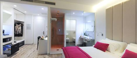 A modern hotel room with a large bed, a walk-in closet, a bathroom with a shower, and a desk with a chair.