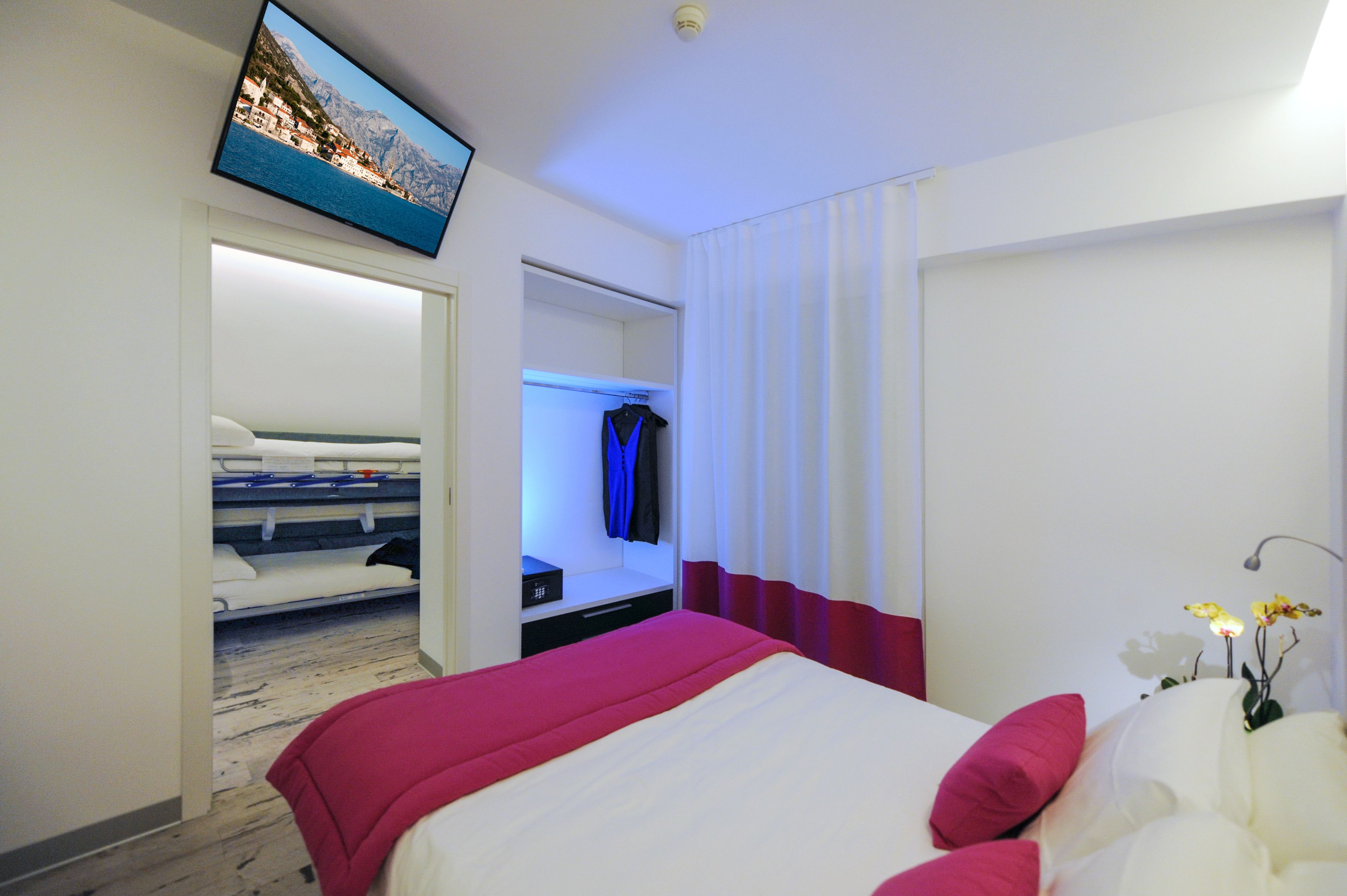 Una habitación de hotel con cama, televisión montada en la pared, armario y ventana con cortinas.