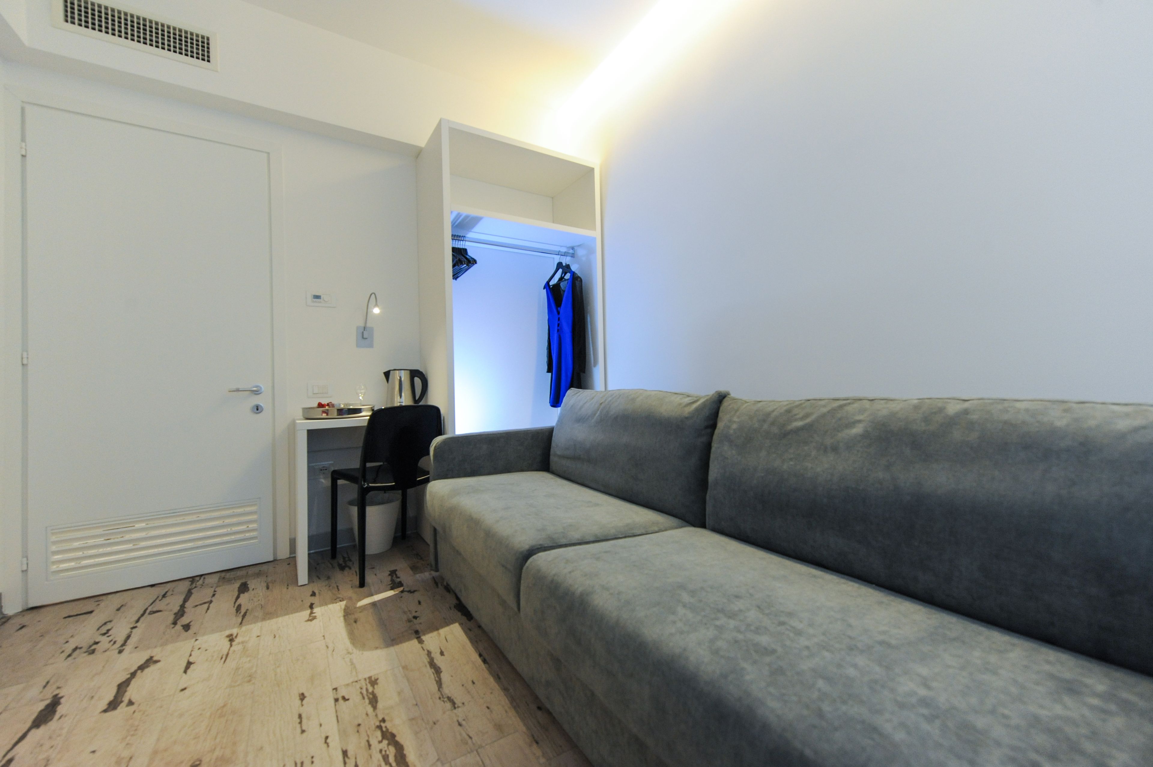 A compact room with a grey sofa, a desk with a chair, and a wardrobe with clothes hanging.