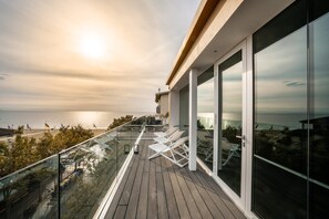 Uma varanda com vista para o mar, cadeiras de lounge brancas e guarda-corpo de vidro.
