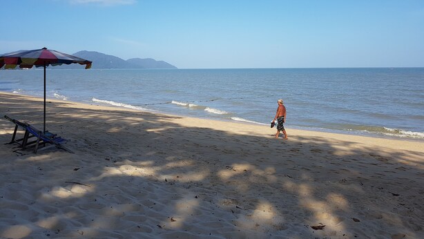A beach with a single umbrella and a chair, a person walking, and a mountain in the background. - D'Feringghi Haus Hotel & Restaurant (George Town)