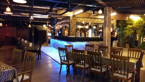 A restaurant with wooden chairs and tables, a bar area with decorative lights, and a stone wall backdrop. - D'Feringghi Haus Hotel & Restaurant (George Town)