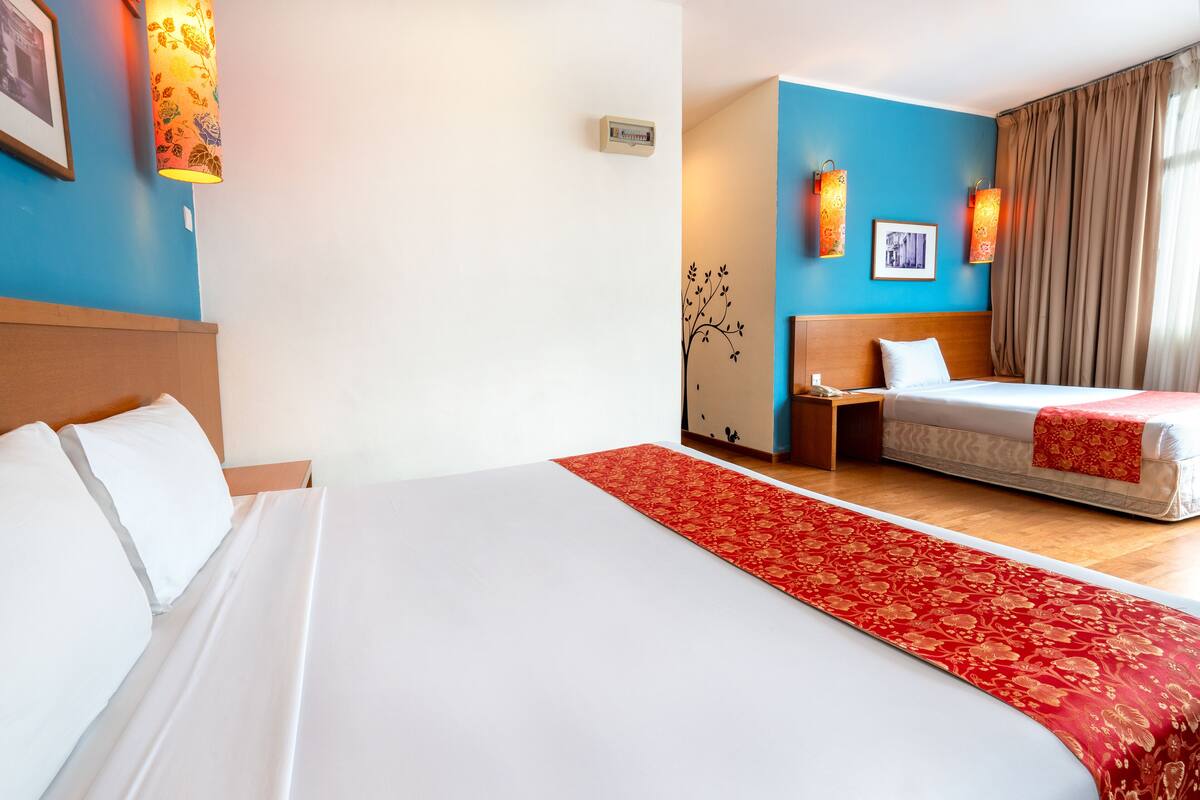 Una camera d'albergo con un letto grande, un letto più piccolo, un tappeto decorativo rosso, una lampada a parete e un portafoto.