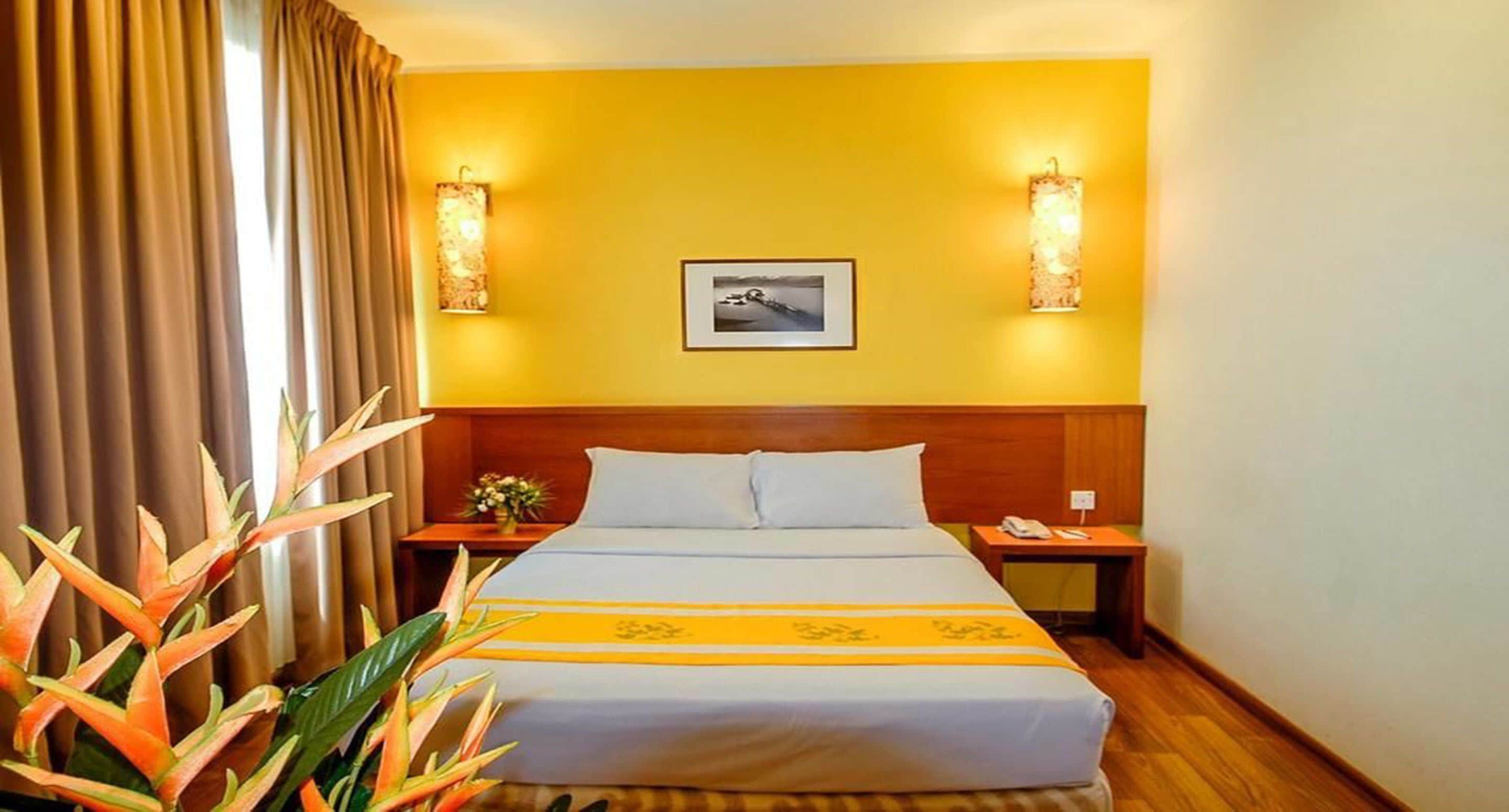 Una habitación de hotel con cama, mesita de noche, lámpara, un cuadro en la pared y una planta.