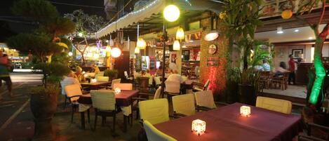 Un restaurant en plein air, doté de tables et de chaises, éclairé par des lampes suspendues et entouré de plantes en pot.
