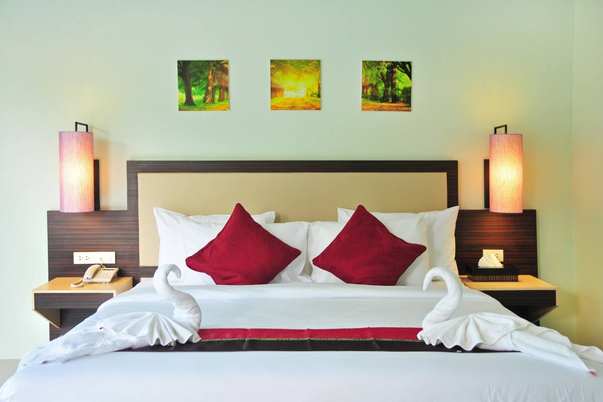 Quarto de hotel com uma cama grande, almofadas vermelhas, duas luminárias fixadas na parede e três quadros enquadrados.