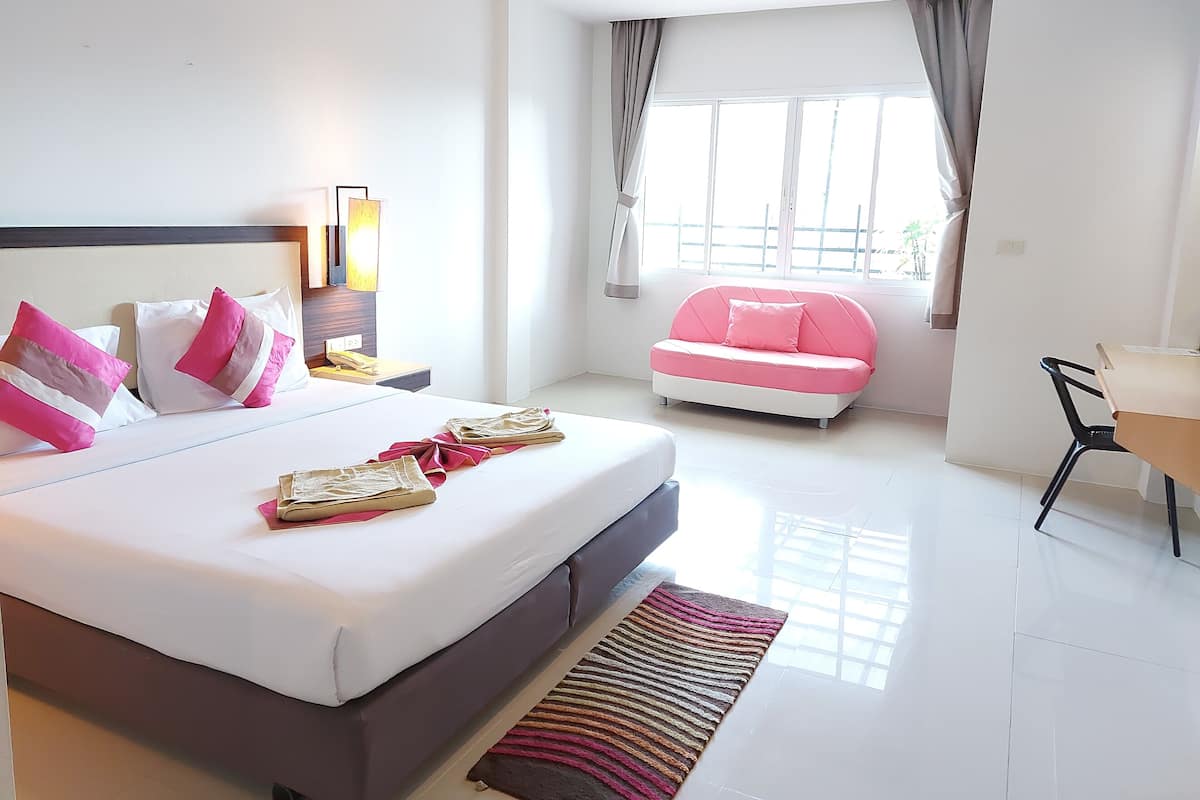Quarto de hotel moderno com uma cama grande, um sofá rosa, uma escrivaninha e uma janela com cortinas.