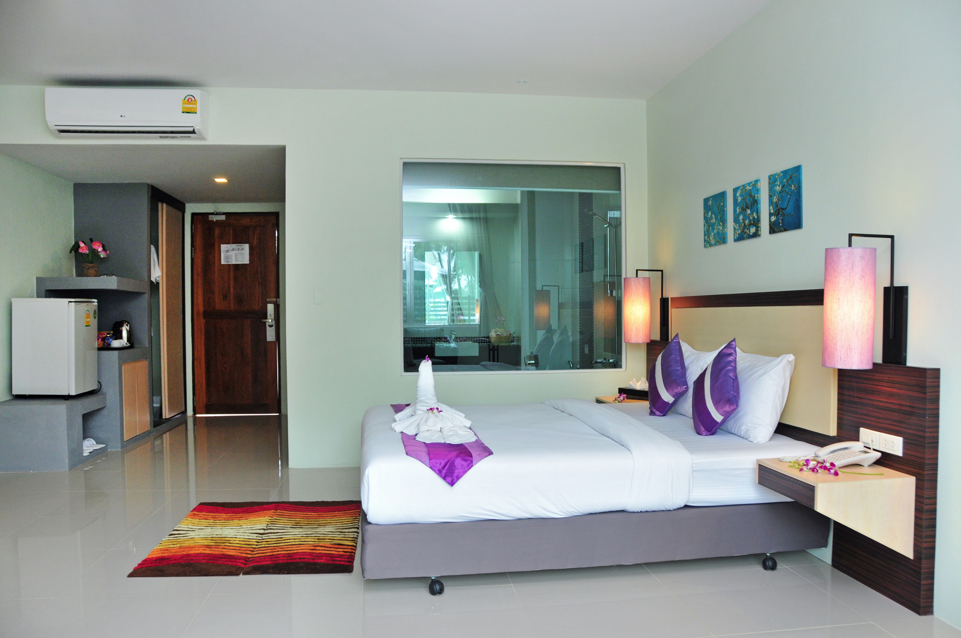 A hotel room with a bed, bedside table, lamp, and a view of the hallway and another room.