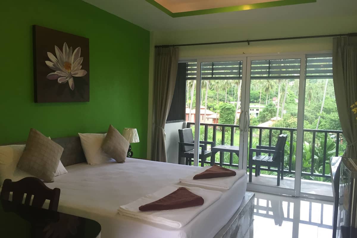 Habitación de hotel con una cama grande, un cuadro de una flor, un balcón con vistas a la vegetación y una mesa con sillas.