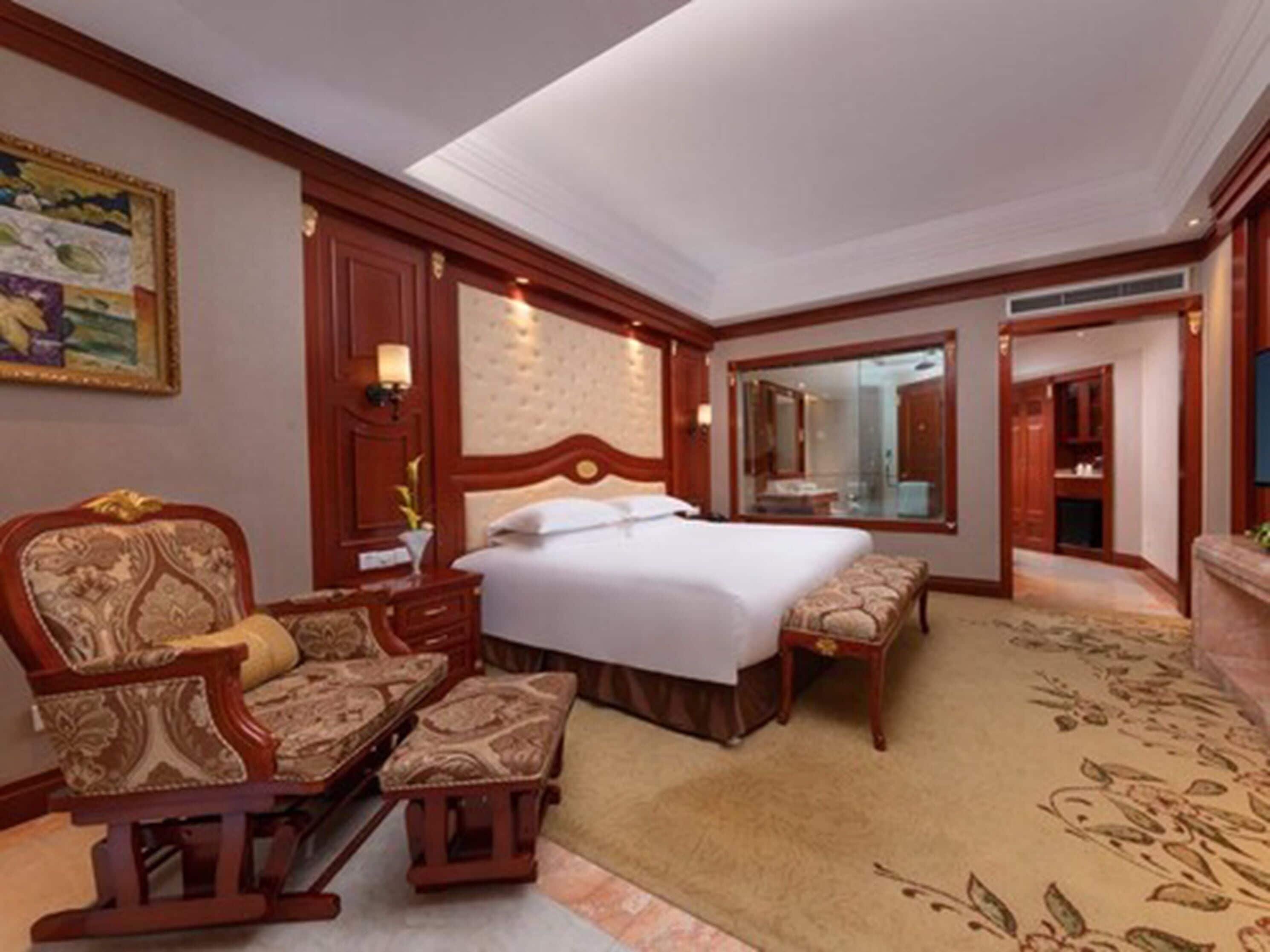 Une chambre d’hôtel dotée d’un grand lit, d’un coin salon avec un fauteuil et un pouf, d’un miroir et d’un sol recouvert de moquette.
