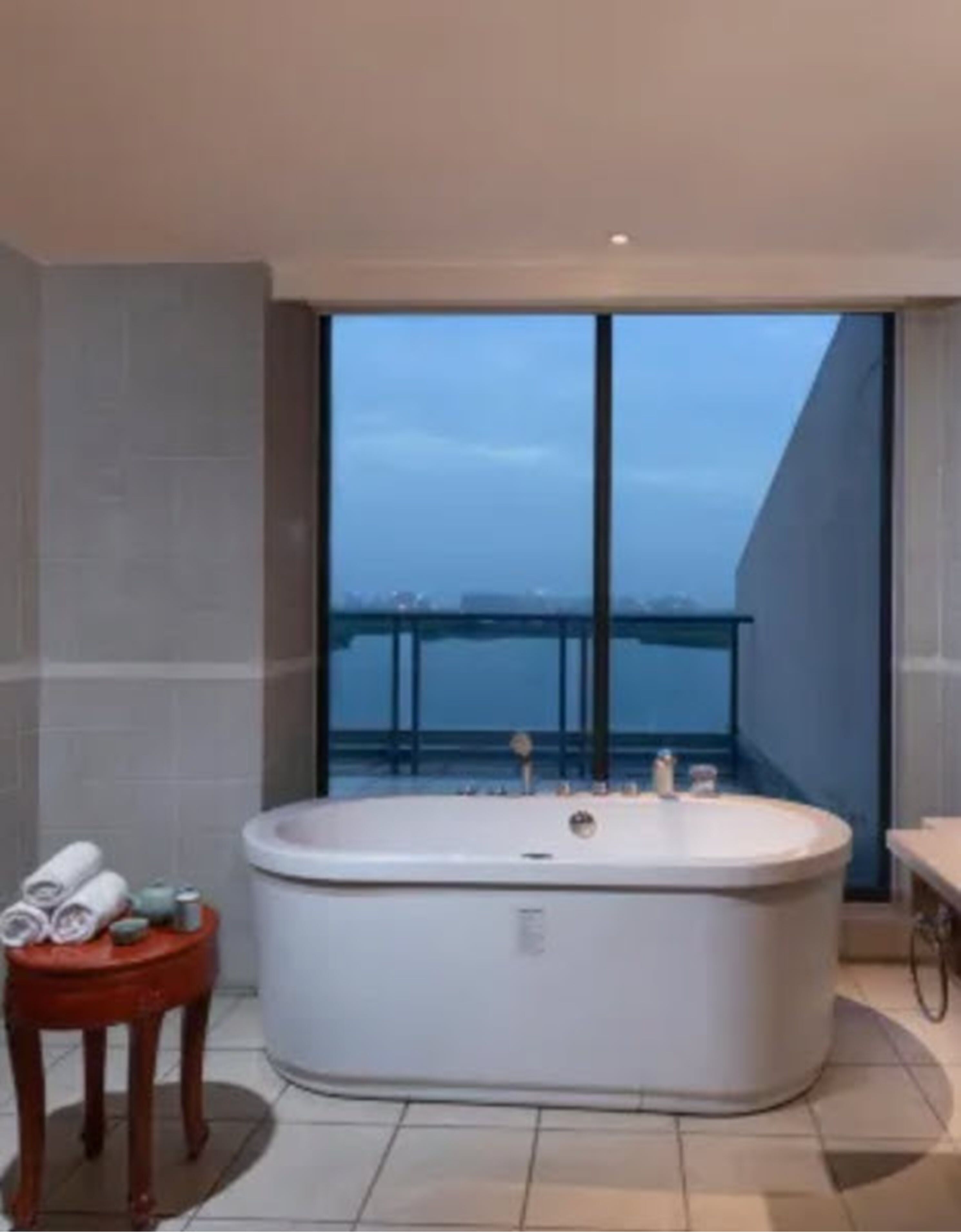Un baño moderno con una bañera independiente, una pequeña mesita de madera con toallas y un ventanal que ofrece vistas a un horizonte urbano.