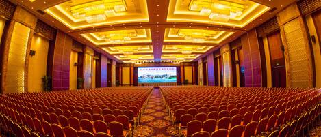 A large conference room with rows of red chairs, a central stage, and ornate ceiling lights.