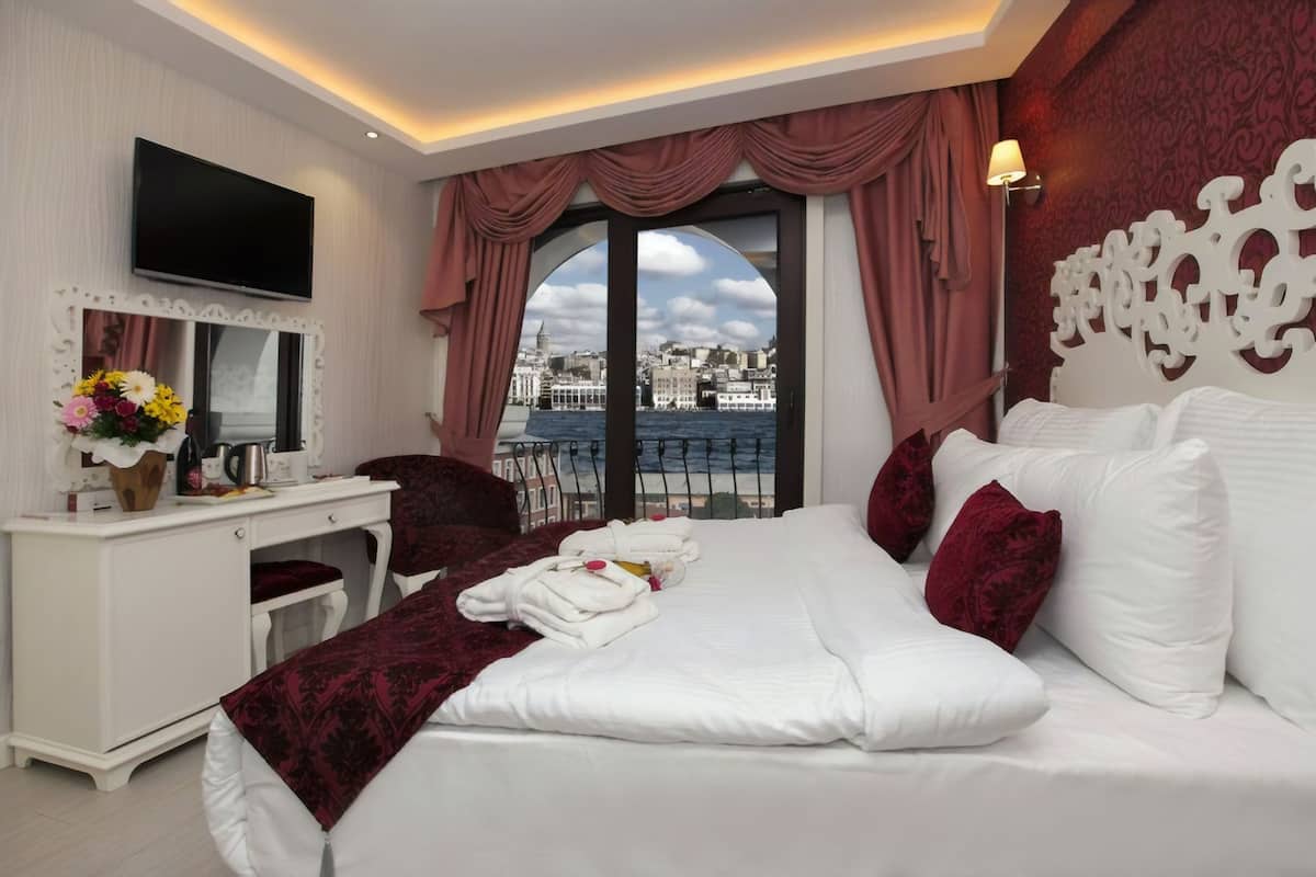 A hotel room with a bed, a desk, a TV, and a view of the city through large windows.