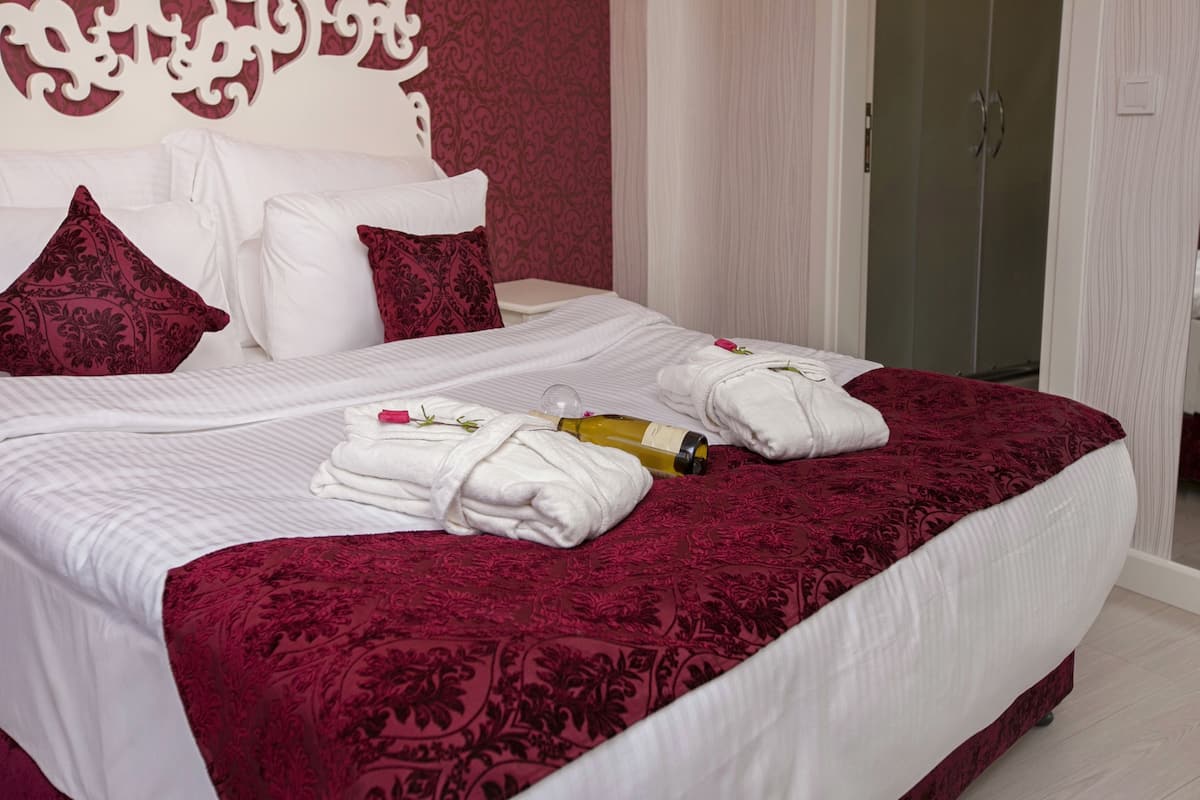 A neatly made bed with white linens, a red patterned bedspread, and a bottle of champagne.