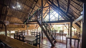 Un espace de restauration intérieur avec des tables et des bancs en bois, un escalier et une structure dotée d’un toit de chaume.