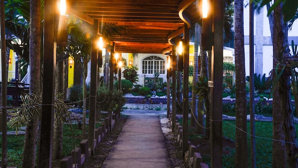 Une allée couverte, agrémentée de piliers en bois et de guirlandes lumineuses, mène à un bâtiment doté d’une grande fenêtre et d’une porte.