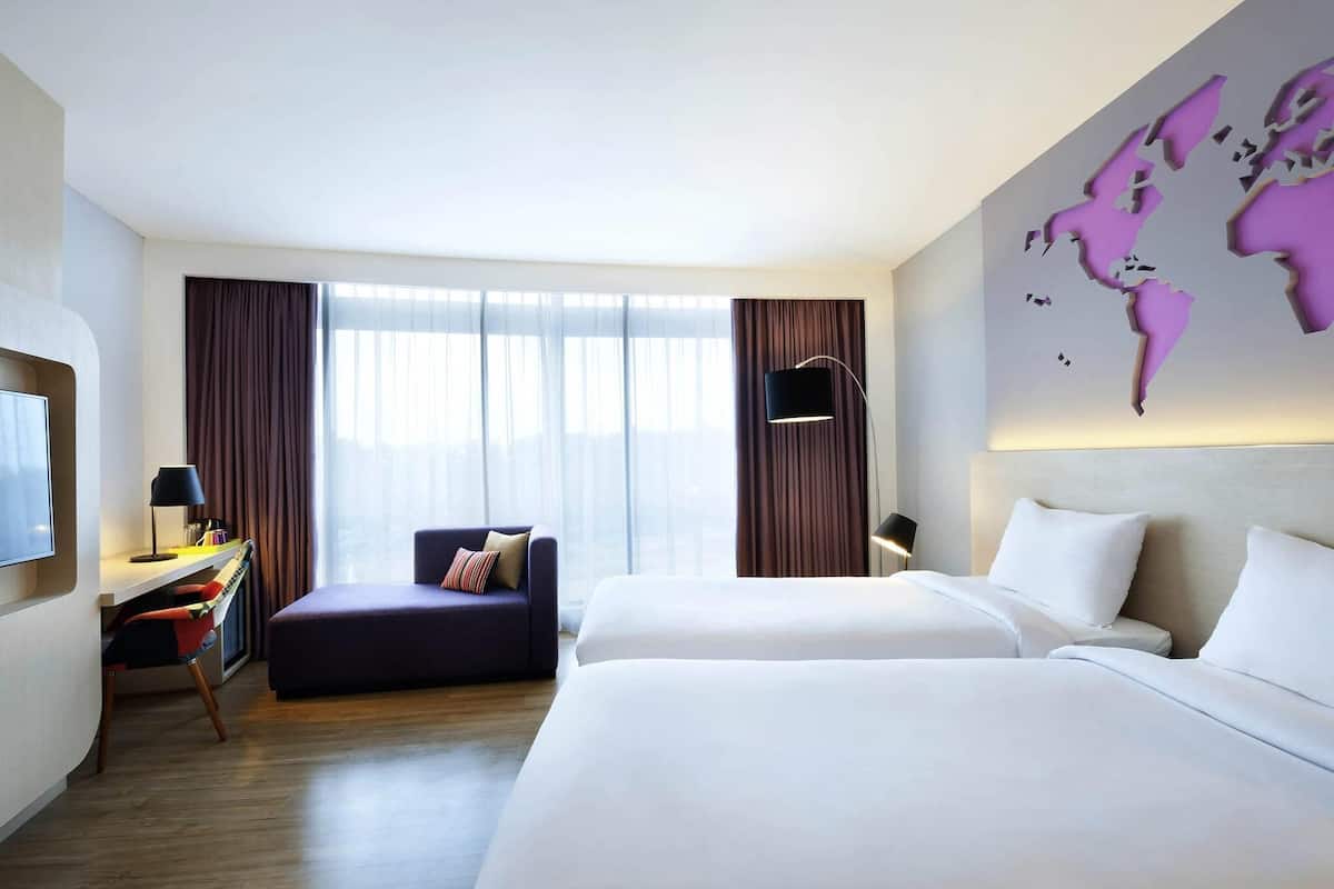 A hotel room with a bed, a desk with a chair, a sofa, a world map on the wall, and a large window with curtains.