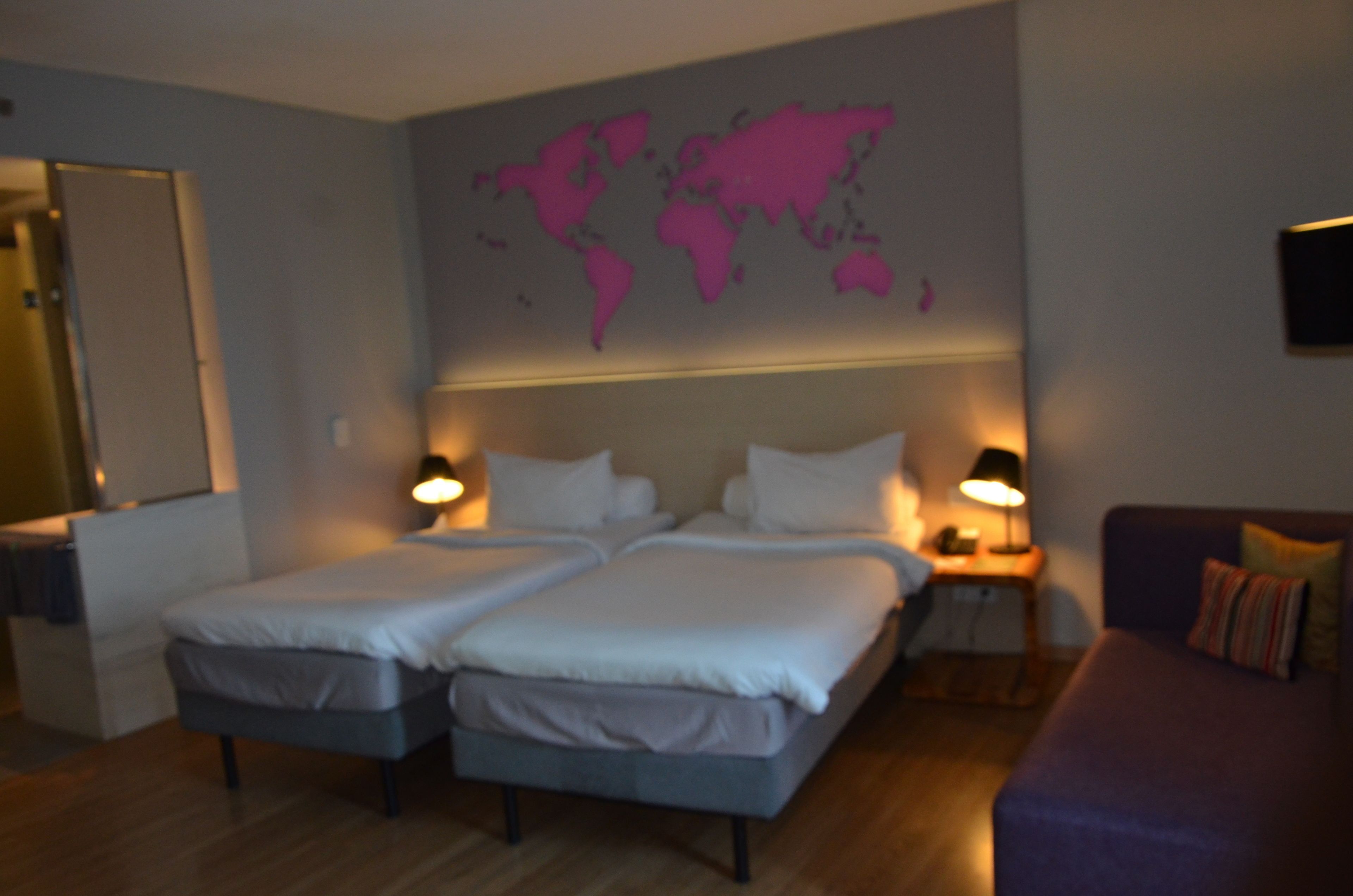 Un lit double avec deux oreillers, une table de chevet avec une lampe, une carte du monde accrochée au mur et un canapé violet.