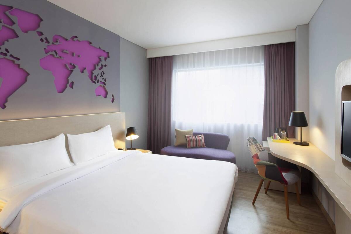 A hotel room with a large bed, a desk, a chair, a world map on the wall, and a window with curtains.