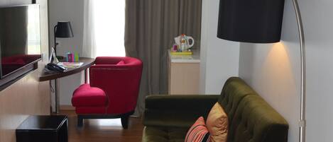 Uma sala de estar moderna com um sofá verde, uma poltrona vermelha, uma TV de tela plana, uma escrivaninha e um abajur.
