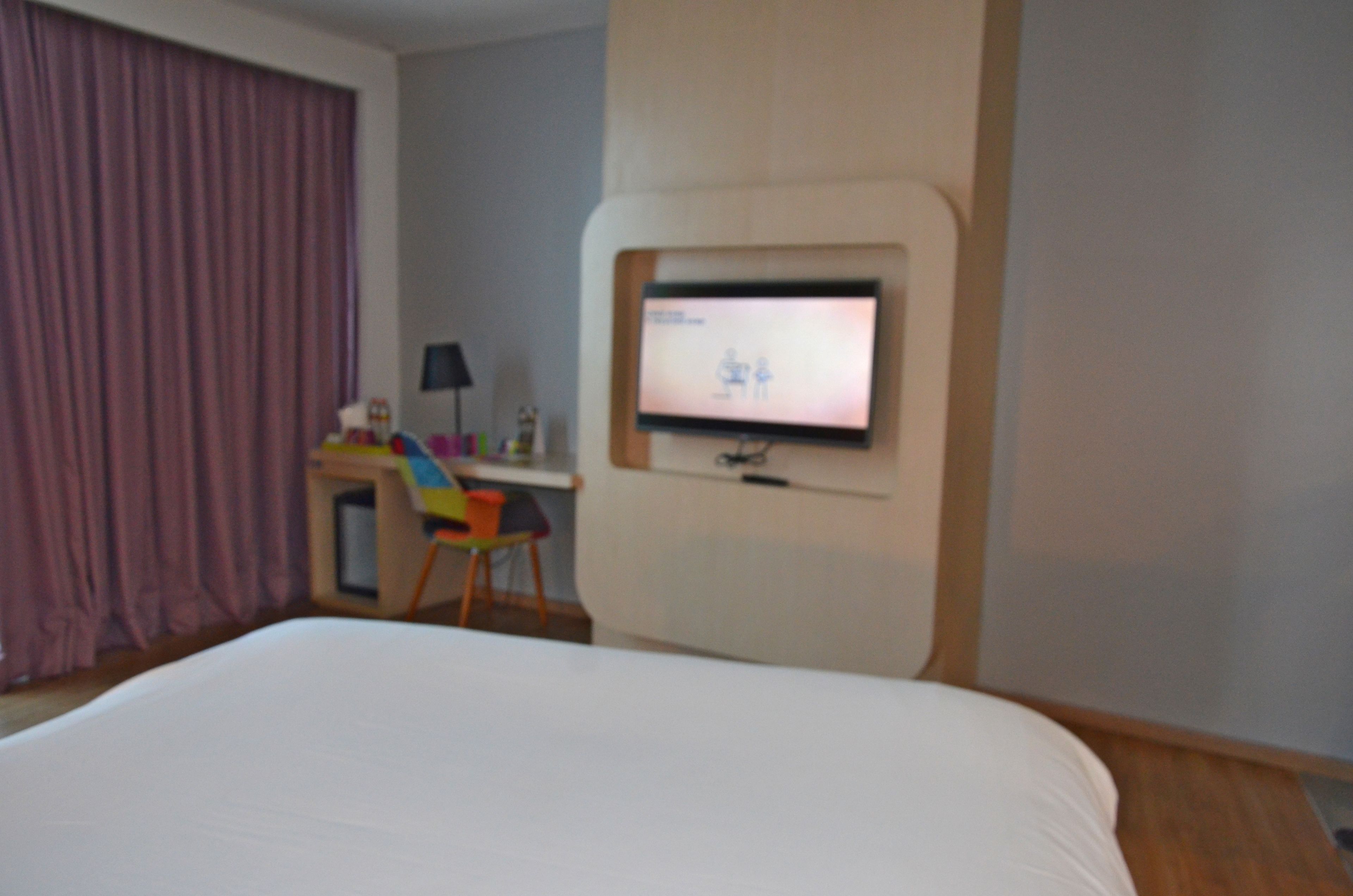 A hotel room with a bed, a desk with a computer, and a television mounted on the wall.