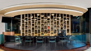 A bar area with a curved wooden counter, multiple shelves with bottles, and a seating area with chairs. - ibis Styles Jakarta Airport (Tangerang)