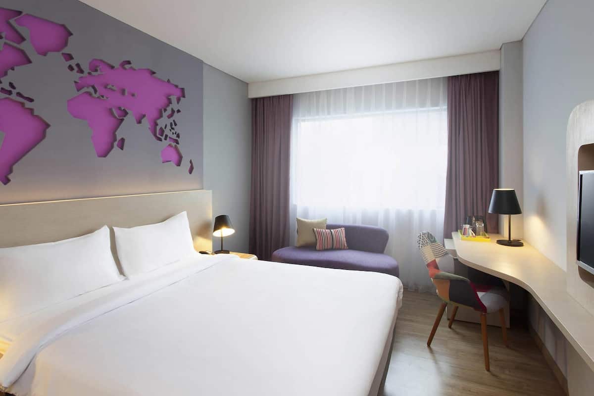 A hotel room with a large bed, a desk, a chair, a world map on the wall, and a window with curtains.