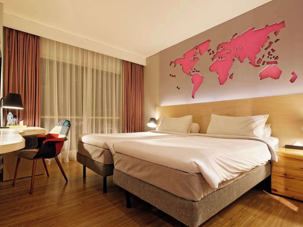 A hotel room with a large bed, a desk with a chair, a world map on the wall, and a window with curtains.