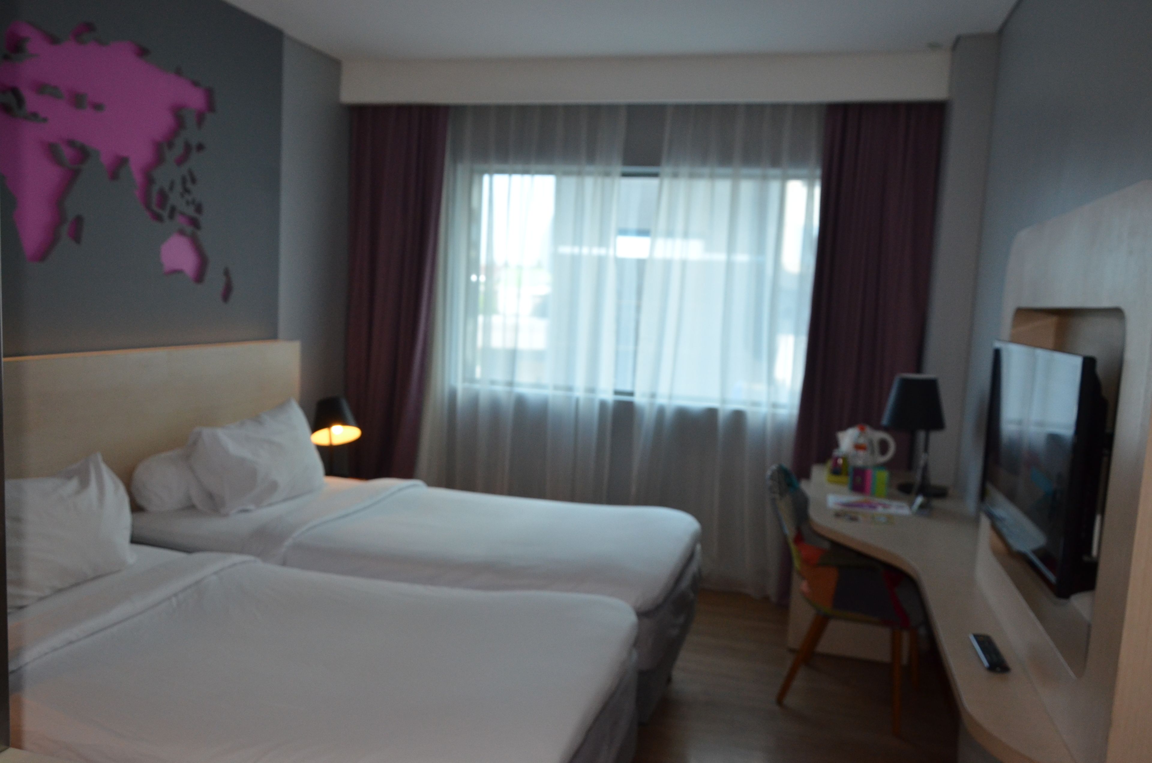Habitación de hotel con dos camas, un escritorio con televisor, un mapa mundial en la pared y una ventana con cortinas.