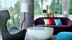 A modern living room with a glass coffee table, a purple sofa with colorful cushions, and a wicker chair. There are two lamps with white shades and a laptop on a glass table. The room has large windows with a view of greenery outside. - ibis Styles Jakarta Airport (Tangerang)