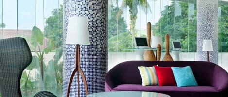 Una sala de estar moderna con una mesa de centro de vidrio, un sofá púrpura con cojines coloridos y una silla de mimbre. Hay dos lámparas con pantallas blancas y un ordenador portátil sobre una mesa de vidrio. La habitación tiene ventanas amplias con vistas a zonas verdes.
