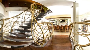 A spiral staircase with a modern design, leading to a dining area with wooden furniture and a bar in the background. - Köstem Hotel (Cesme)