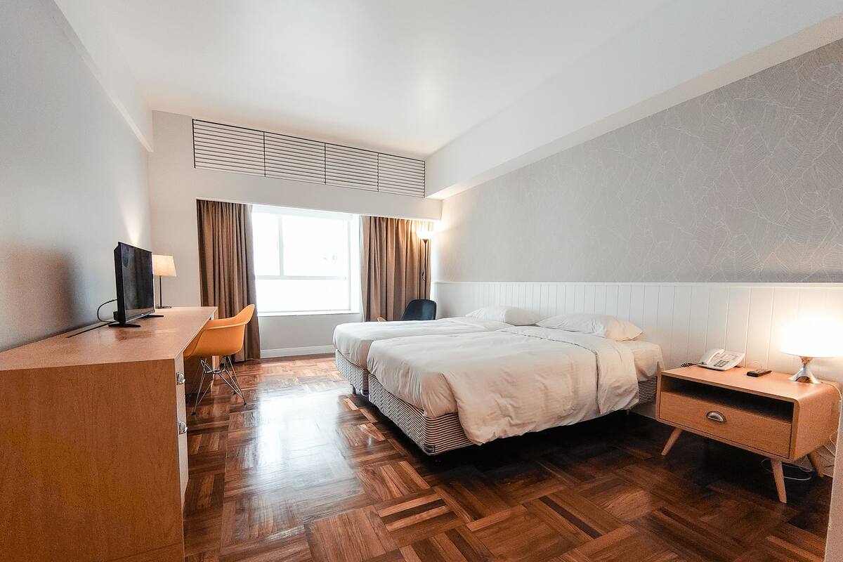 Habitación de hotel con suelo de parqué de madera, cama con ropa de cama blanca, escritorio con computadora, silla, mesita de noche con lámpara y ventana con cortinas.