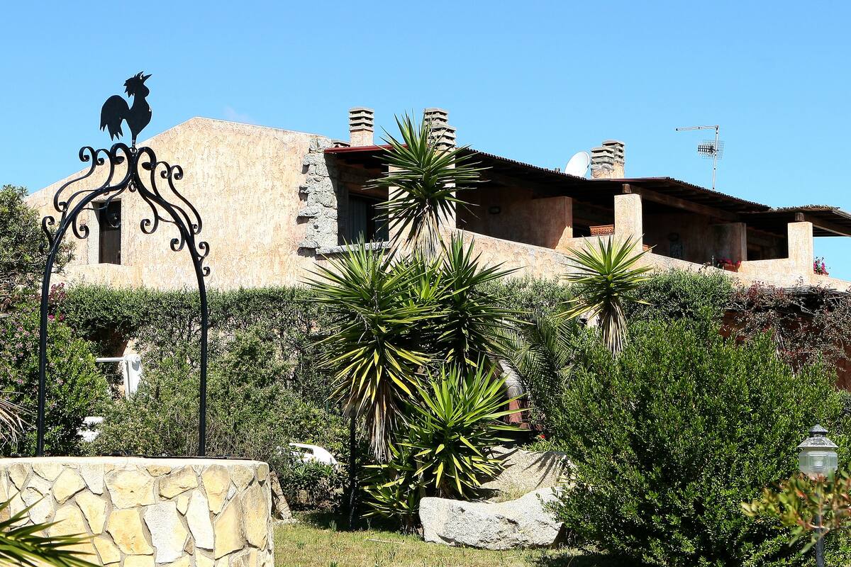 Un bâtiment aux murs en pierre et au toit plat, entouré de verdure et d’une clôture en métal décorative.