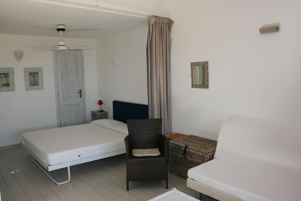 Une pièce comprenant un lit, une chaise, une petite table, un miroir fixé au mur et un ventilateur de plafond.