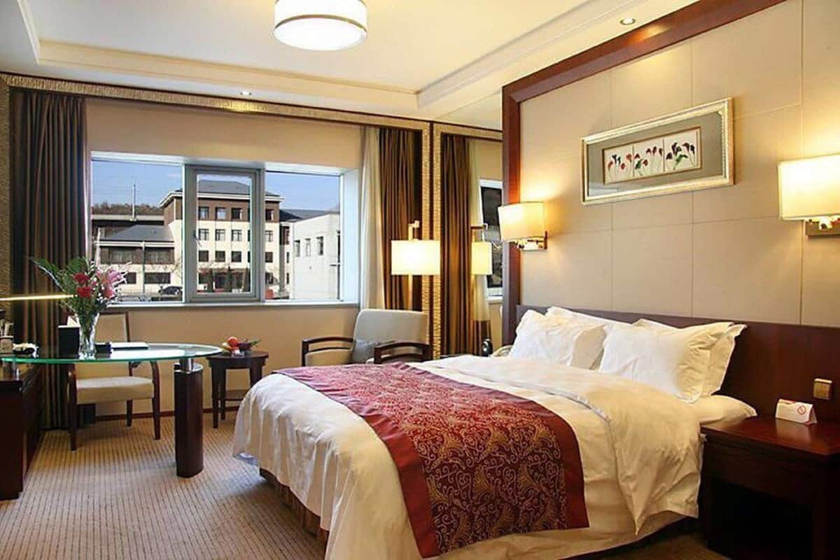 A hotel room with a large bed, a desk with a chair, a window with curtains, and a framed picture on the wall.