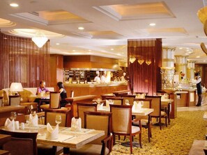 A hotel dining area with multiple tables and chairs, a central bar area, and a kitchen in the background.