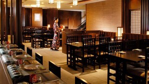 A restaurant with multiple tables and chairs, a person in traditional attire, and a staircase in the background. - Inn Fine Hotel (Dalian)