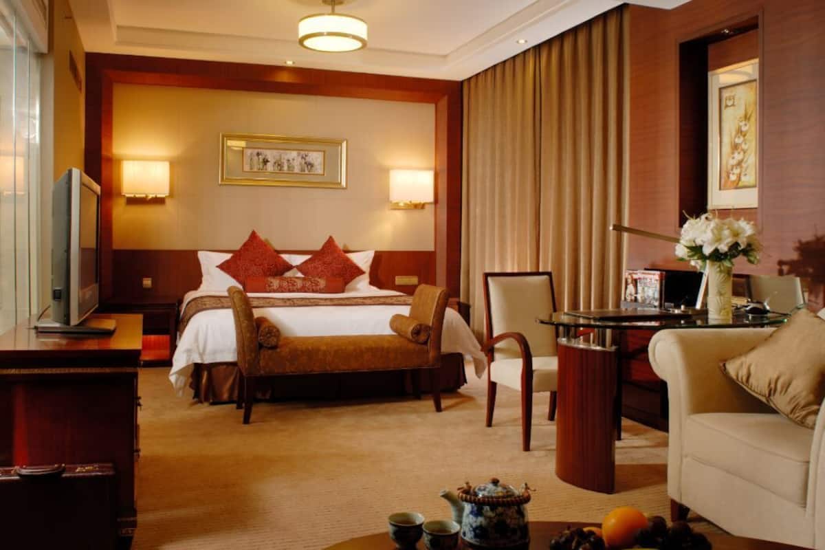 A hotel room with a bed, a desk, a chair, a television, and a coffee table with a teapot and fruit.