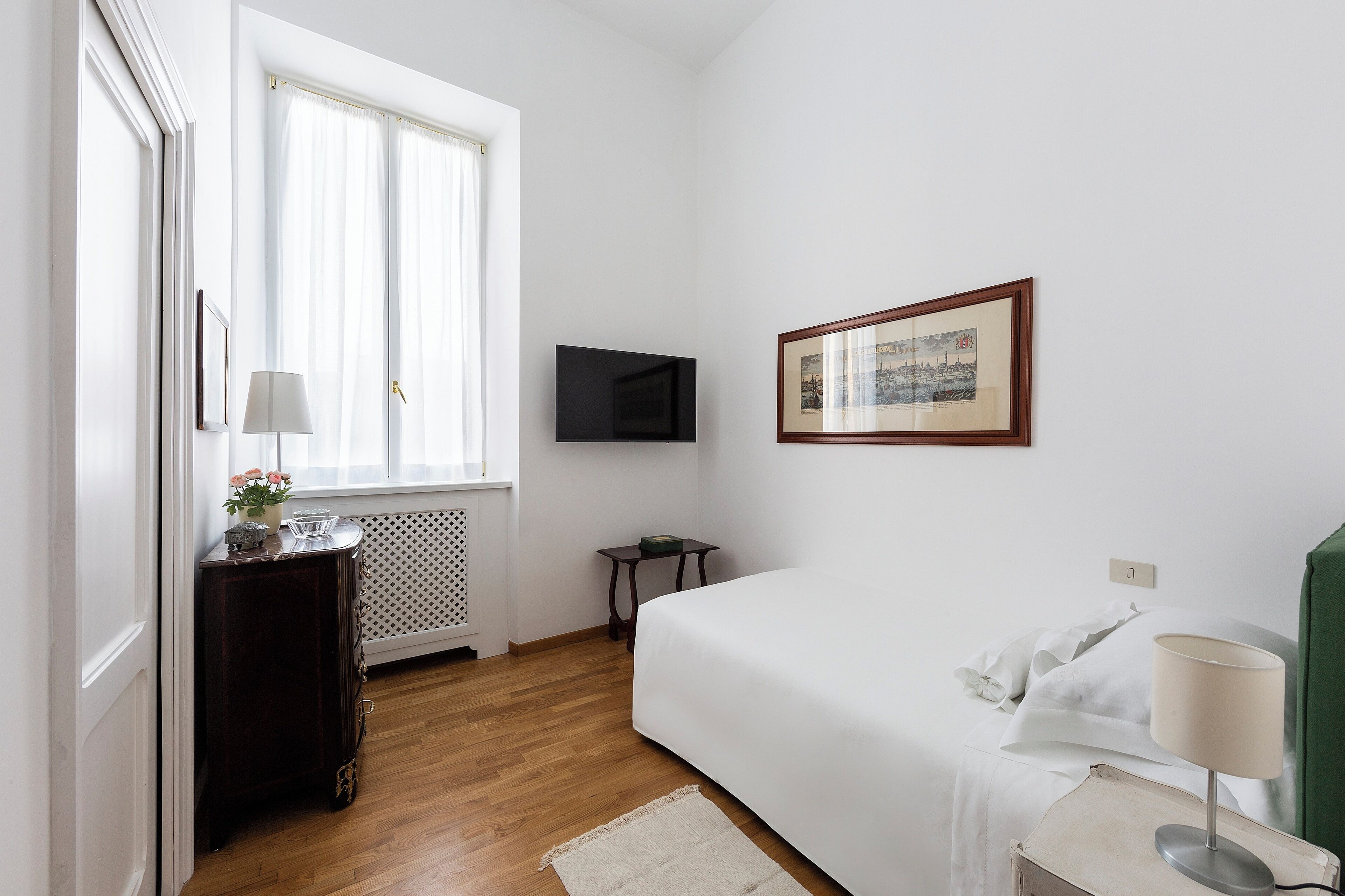 Une chambre à coucher avec un lit, une télévision, un tableau encadré, une commode et une fenêtre avec des rideaux.