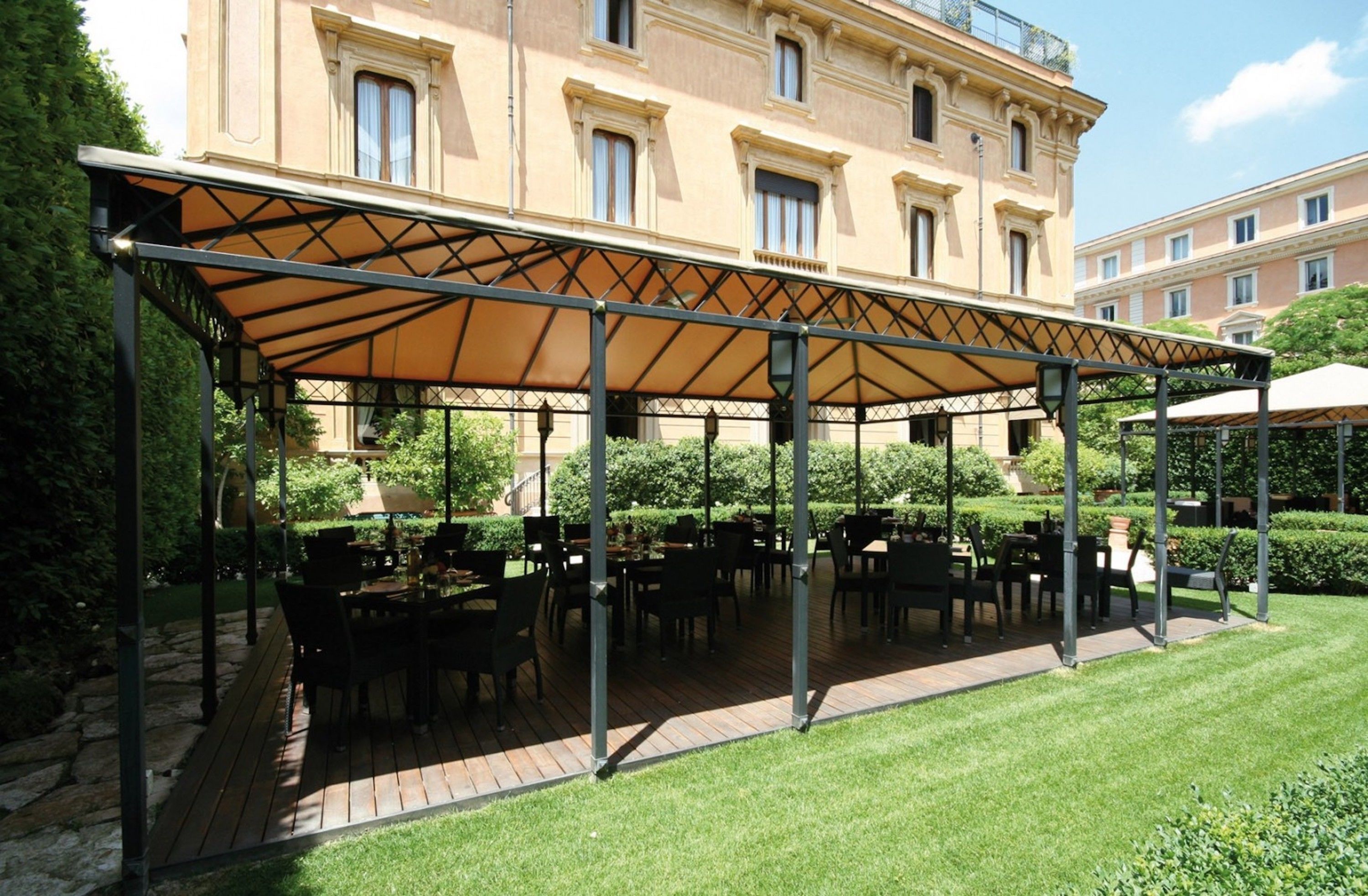 A covered outdoor seating area with tables and chairs, a stone pathway, and a building in the background.