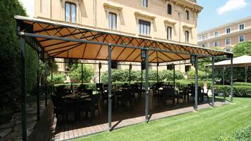 A covered outdoor seating area with tables and chairs, a stone pathway, and a building in the background.