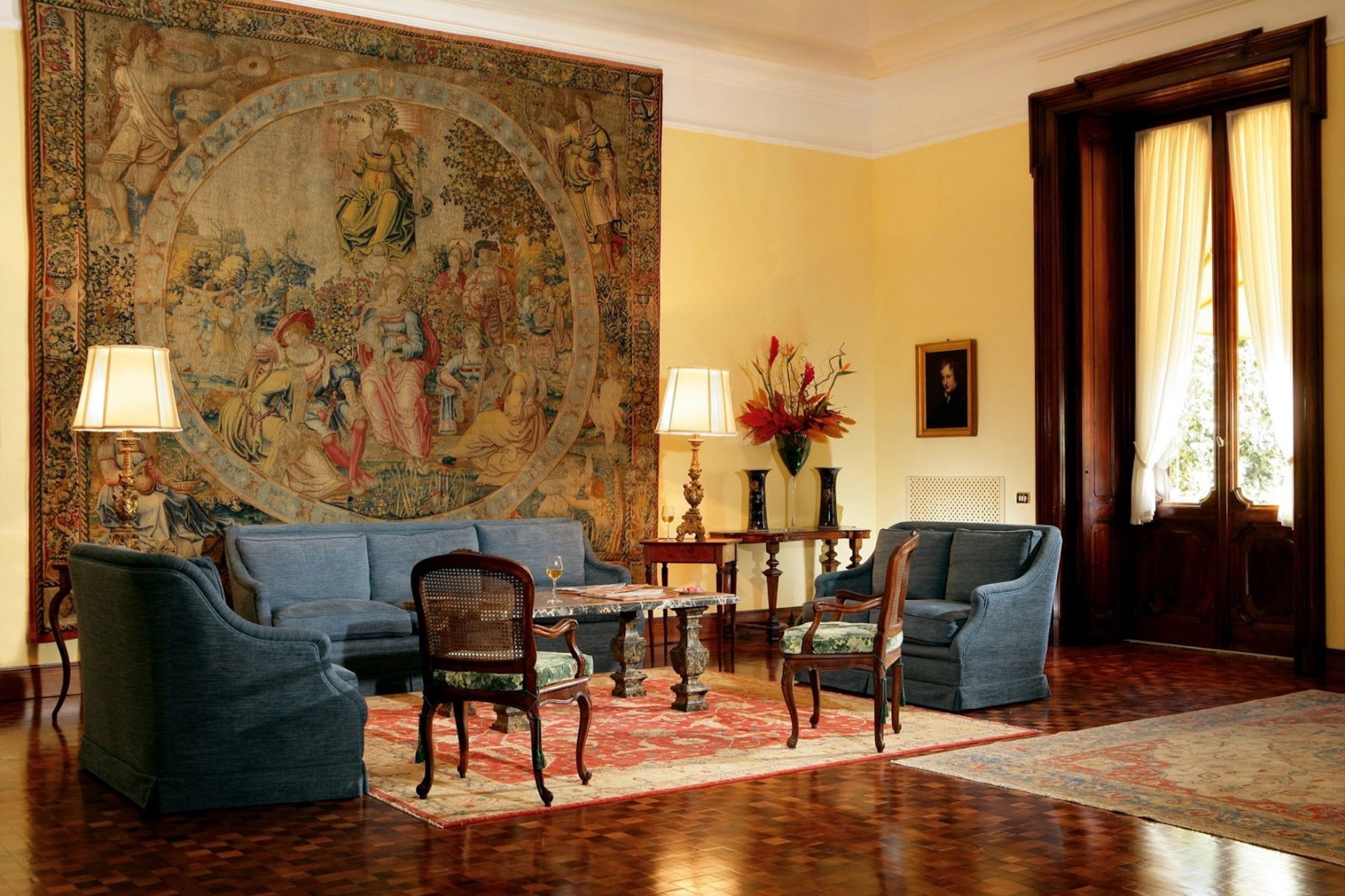 Ruime kamer met een groot wandtapijt, een blauwe bank, twee fauteuils, een klein tafeltje en een houten vloer.