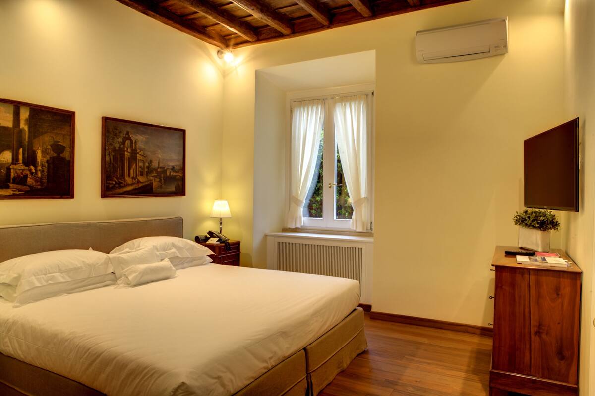 Una camera da letto con un letto, un comodino, una lampada, una finestra con tende, un termosifone, un armadio in legno e due quadri appesi al muro.