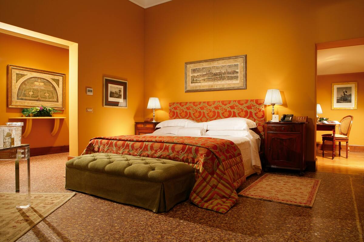 Una camera d'albergo con un letto grande, un poggiapiedi verde, un comodino in legno, una scrivania con una sedia e quadri appesi alle pareti.